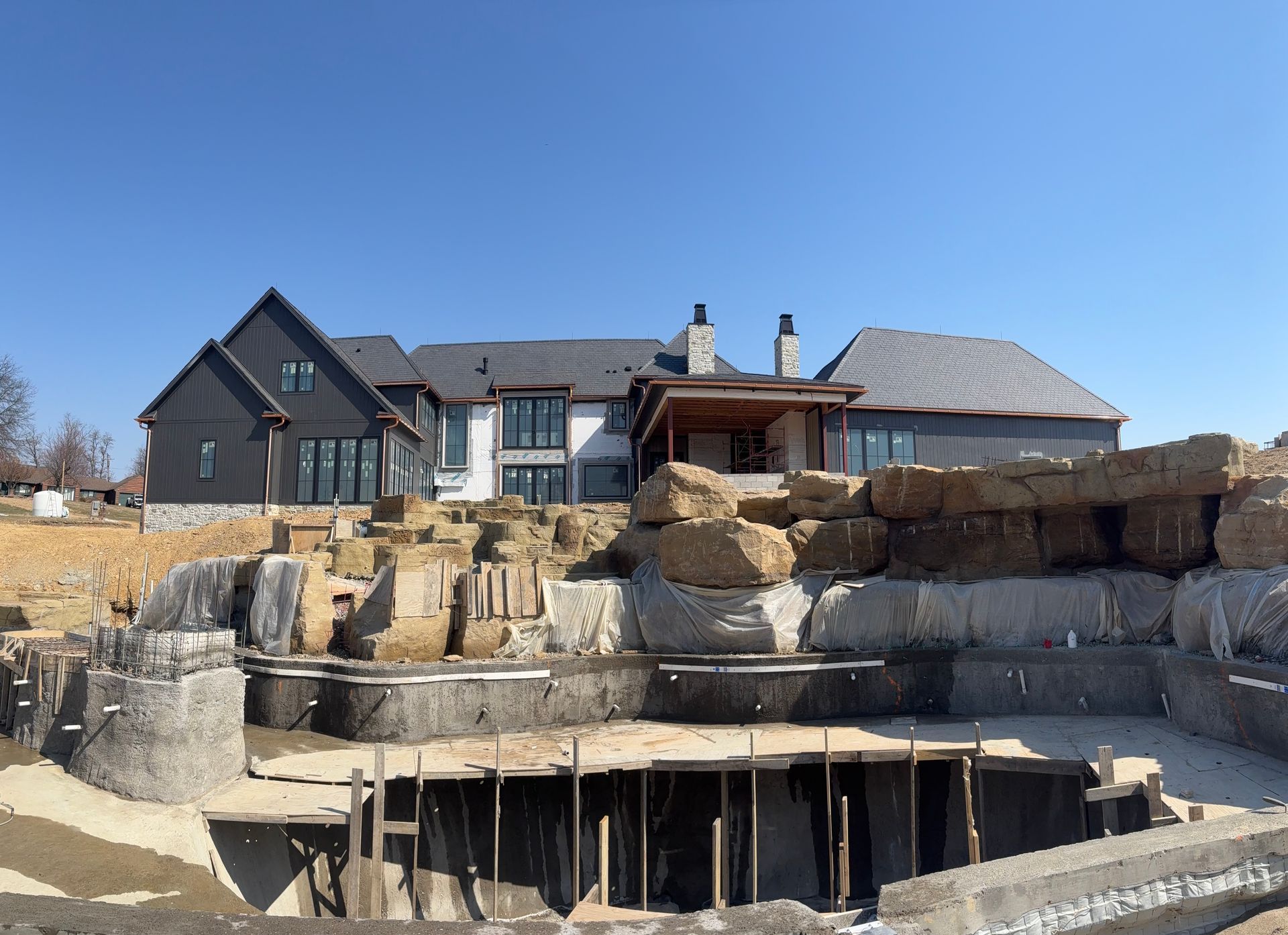 Large modern house under construction beside a rocky excavated foundation under a clear blue sky