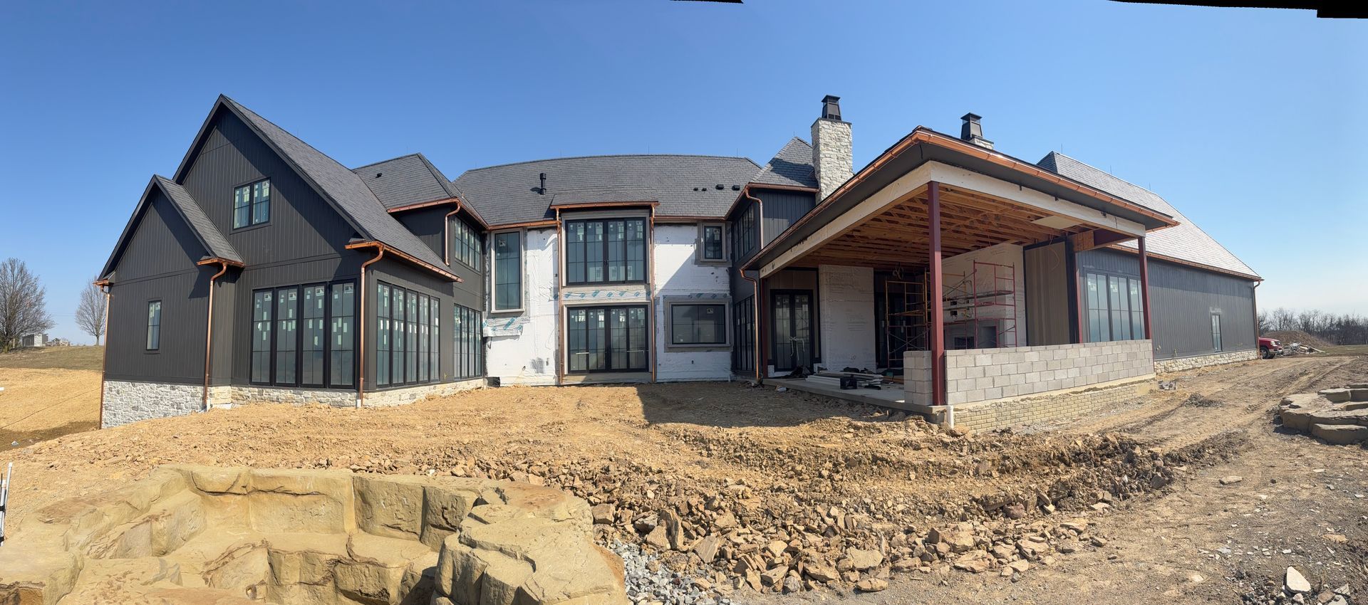 Modern house under construction with dark siding, wood porch, and dirt yard under a clear sky