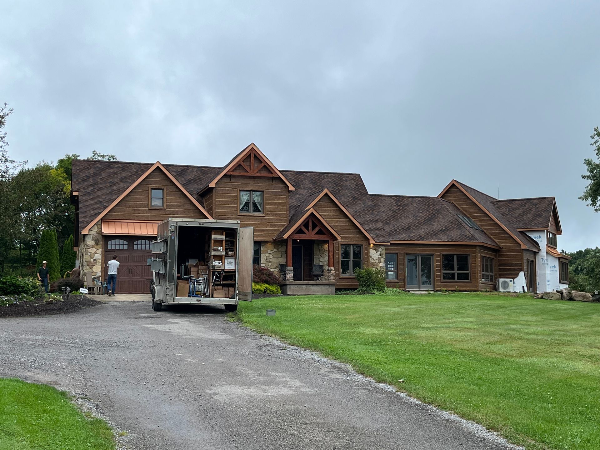 Large brick house with a gravel driveway, green lawn, and parked trailer under a cloudy sky