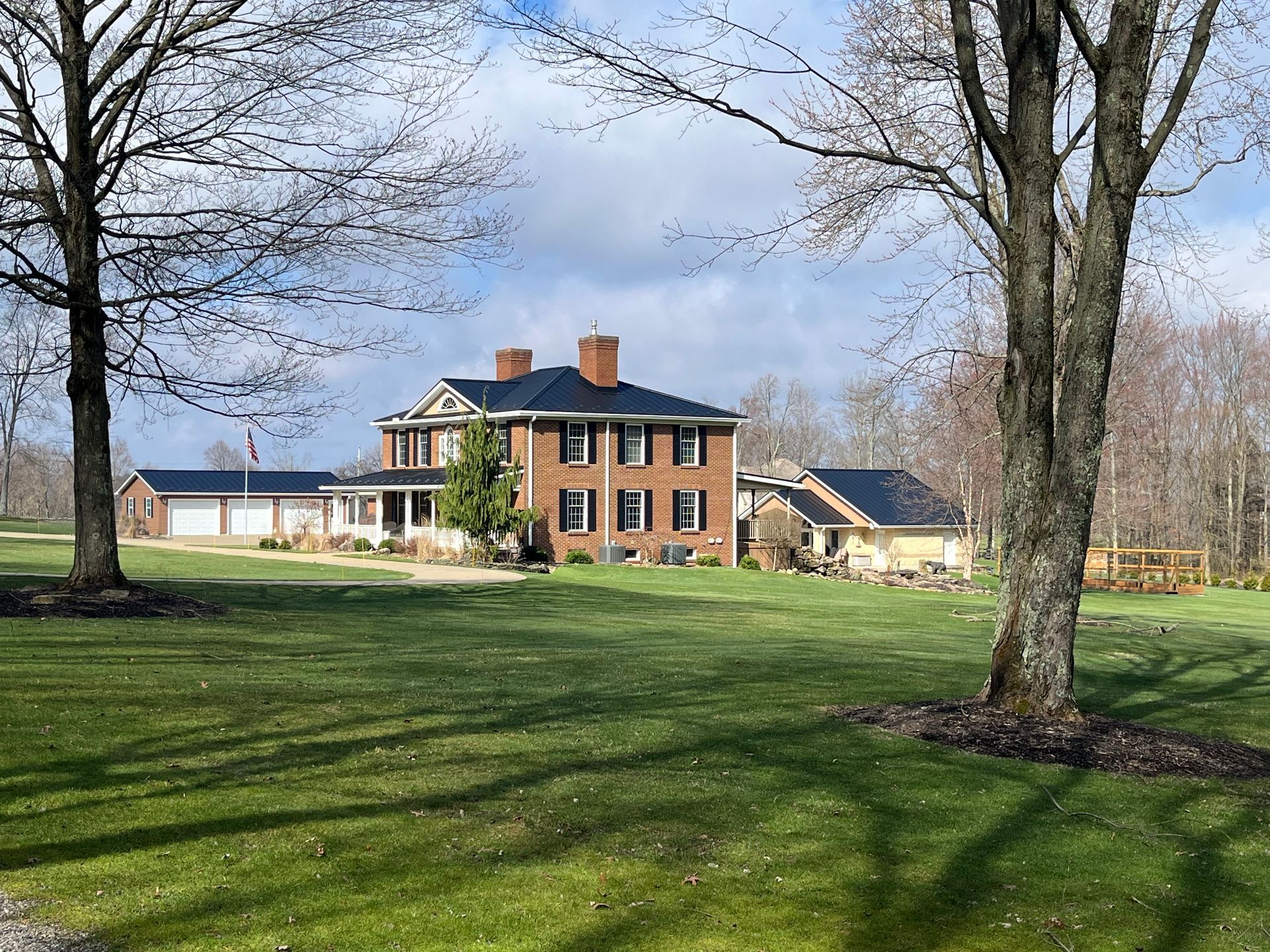 Brick farmhouse with blue roof on a grassy lawn, framed by tall leafless trees under a cloudy sky