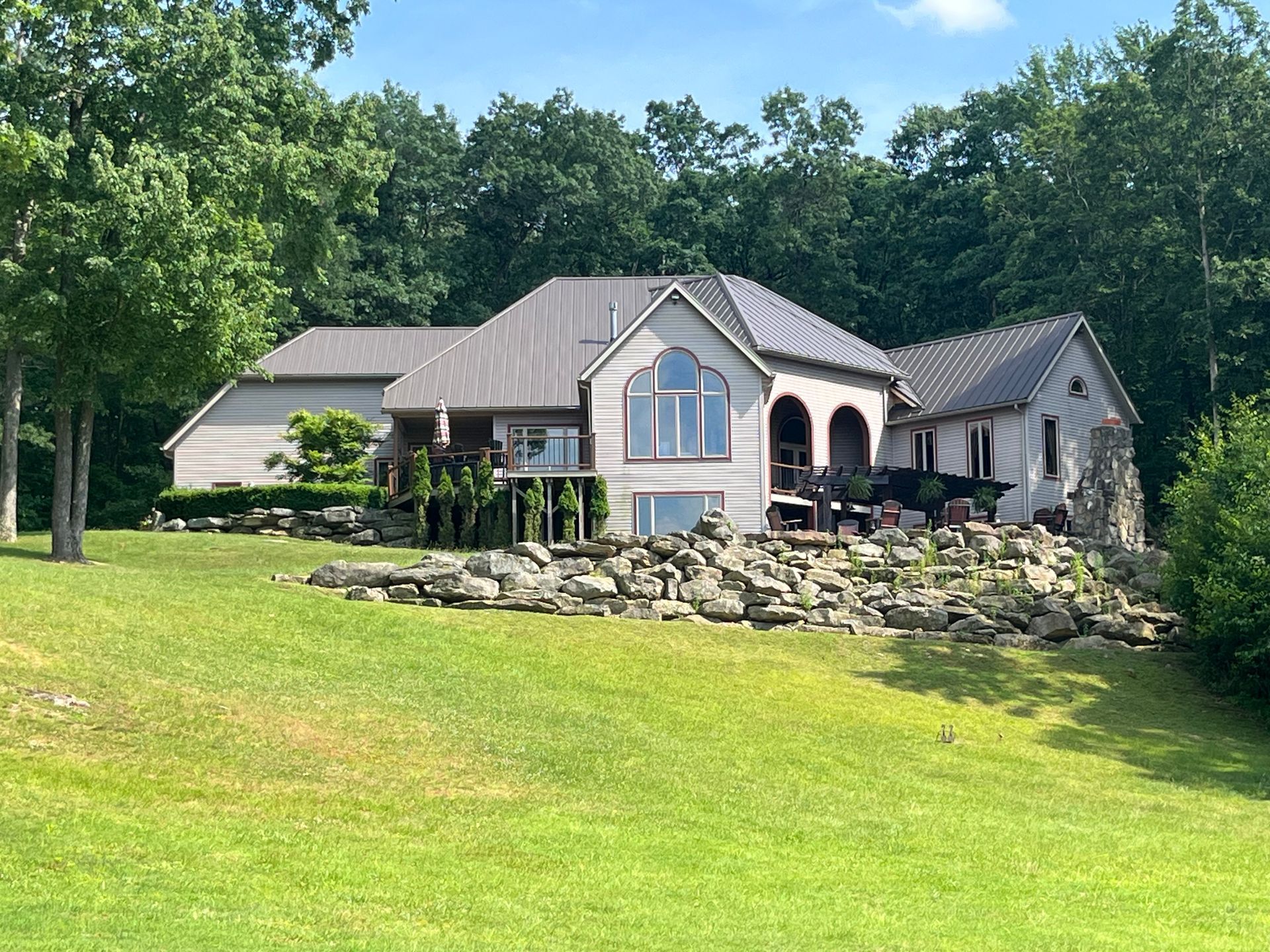 Large gray house on a grassy hillside with a stone retaining wall and trees behind it