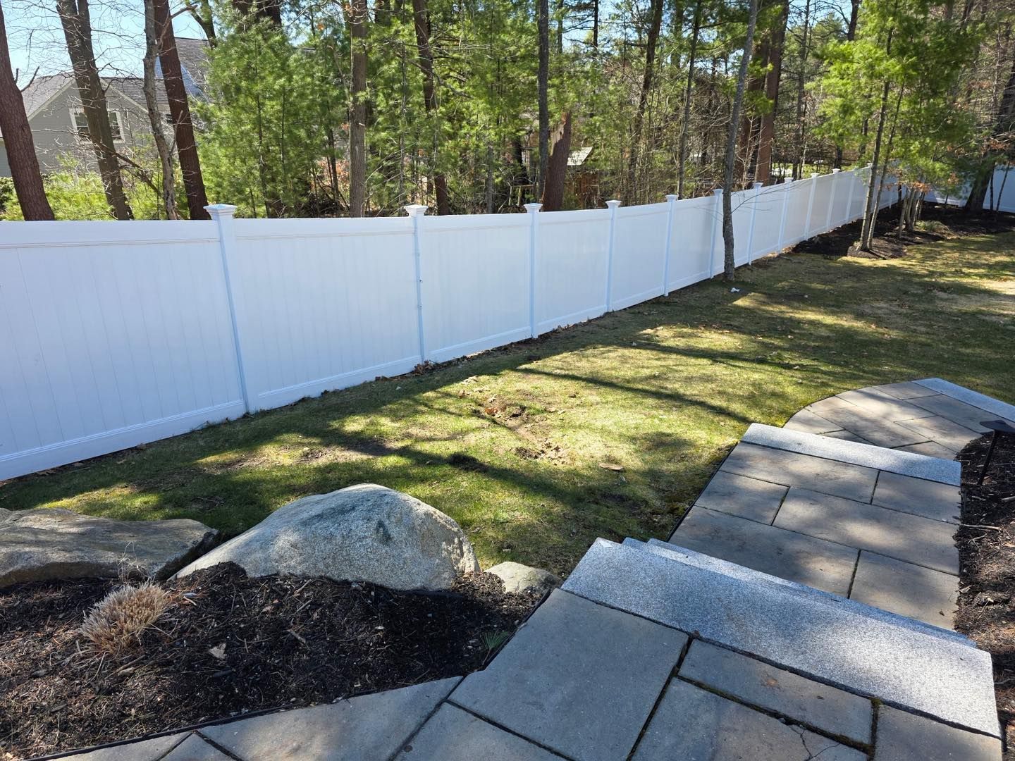 White vinyl fence along a grassy yard beside a paved walkway and trees in sunlight