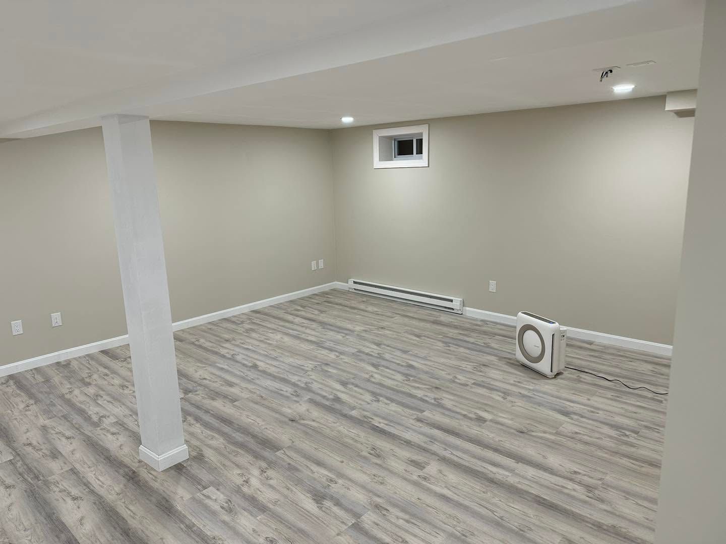 Empty unfinished basement room with beige walls, gray wood-look flooring, and a support column.