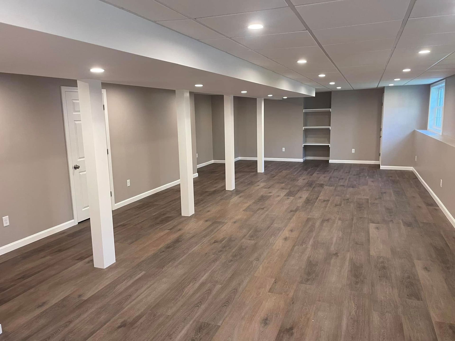 Empty finished basement with gray walls, white support columns, and wood-look flooring.