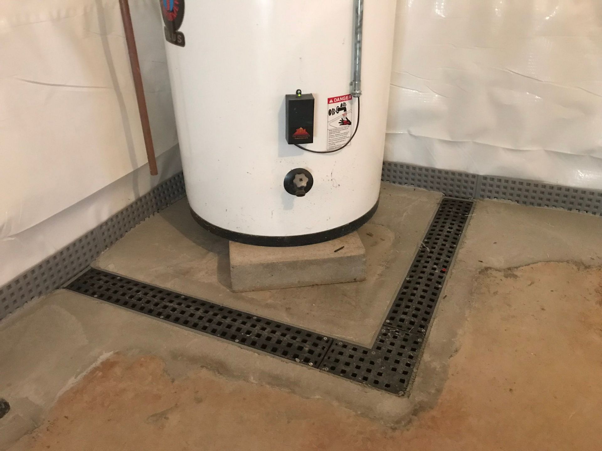 White water heater on a concrete floor with a floor drain and nearby utility piping.