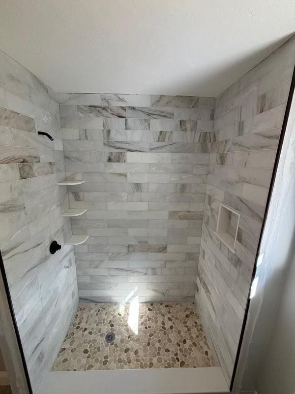 Tiled shower stall with built-in shelves and a pebble-stone floor, lit by natural light.