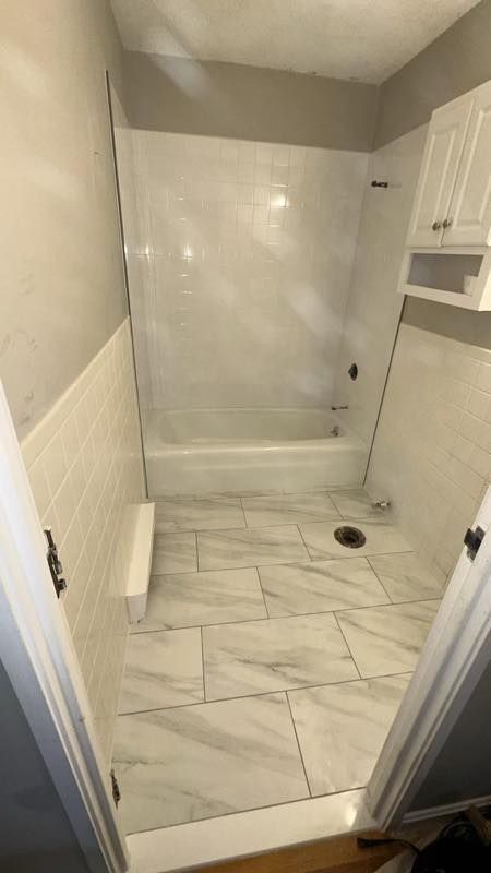 Small white-tiled bathroom with a bathtub-shower combo, marble-look floor, and built-in wall cabinets.