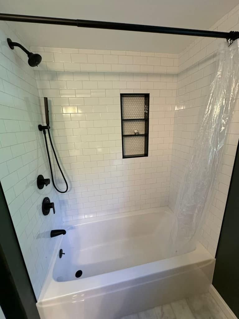 White tiled shower stall with black fixtures, handheld showerhead, built-in tub, and a small frosted window.