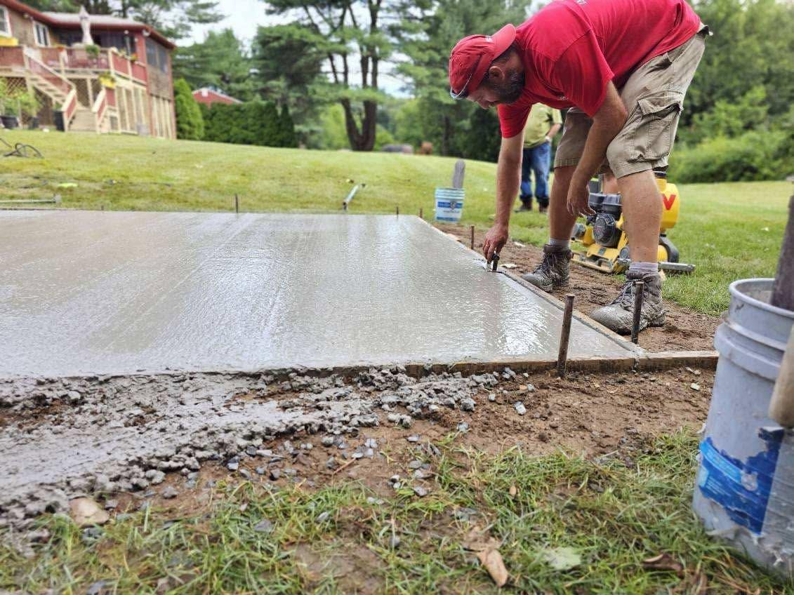 Worker in red shirt smoothing wet concrete on a driveway with tools and buckets nearby
