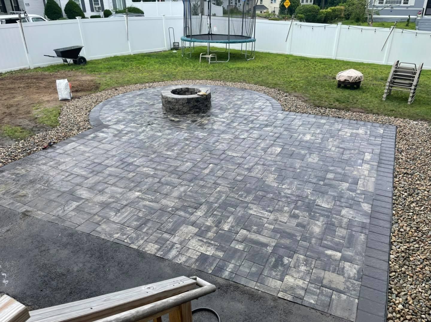Paver patio with a fire pit, surrounded by a grassy yard and a trampoline in the background.