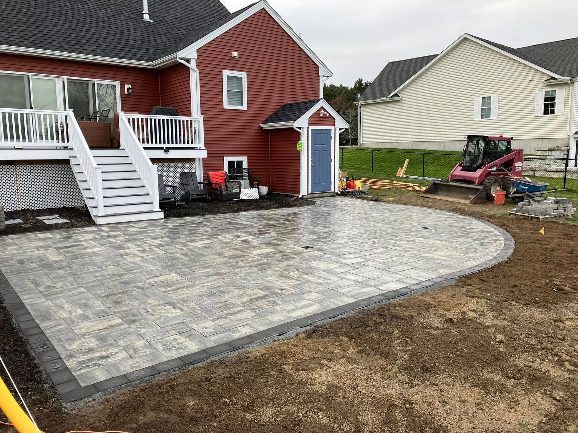 Backyard patio with gray pavers beside a red house, bordered by bare soil and a small shed.