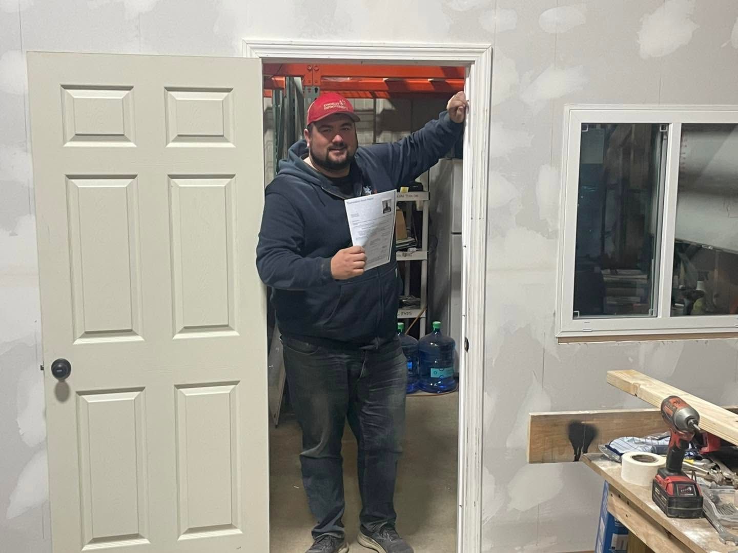 Man standing in a doorway of a room under construction, holding a paper and reaching up near the door frame.