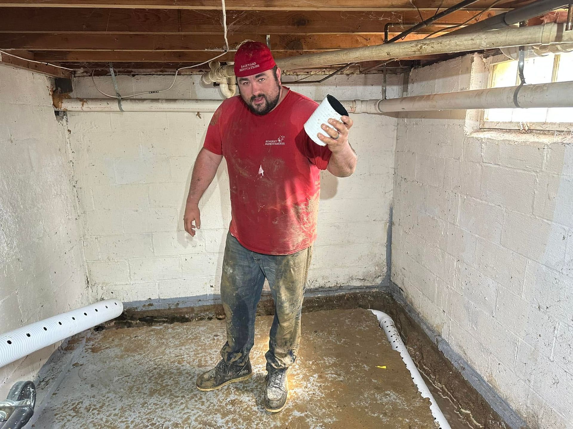 Worker in a red shirt and cap standing in a muddy basement, holding a flashlight.