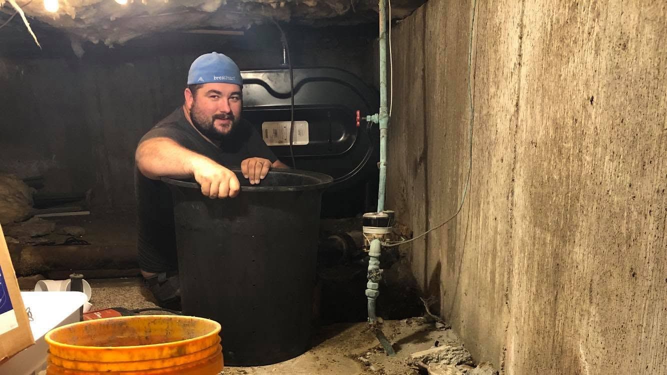 Bearded man in a blue cap leaning on a black barrel in a dim, rustic cellar with stone walls.