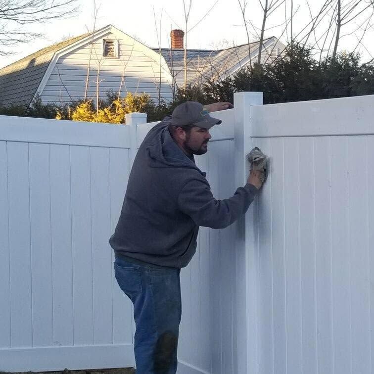 Man painting a white fence panel outdoors with a roller in a suburban yard