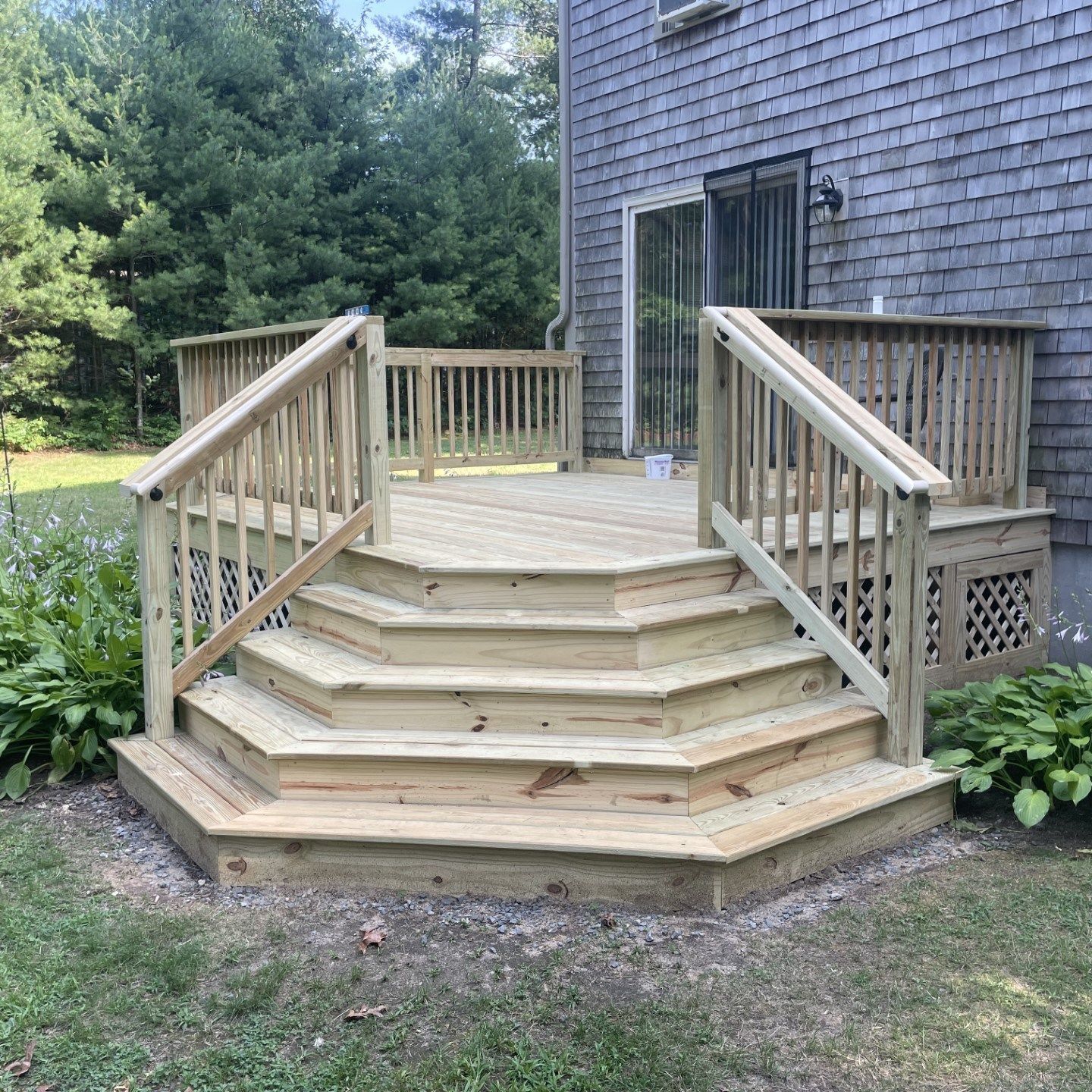 Wooden front steps and deck with railings beside a shingled house, surrounded by greenery