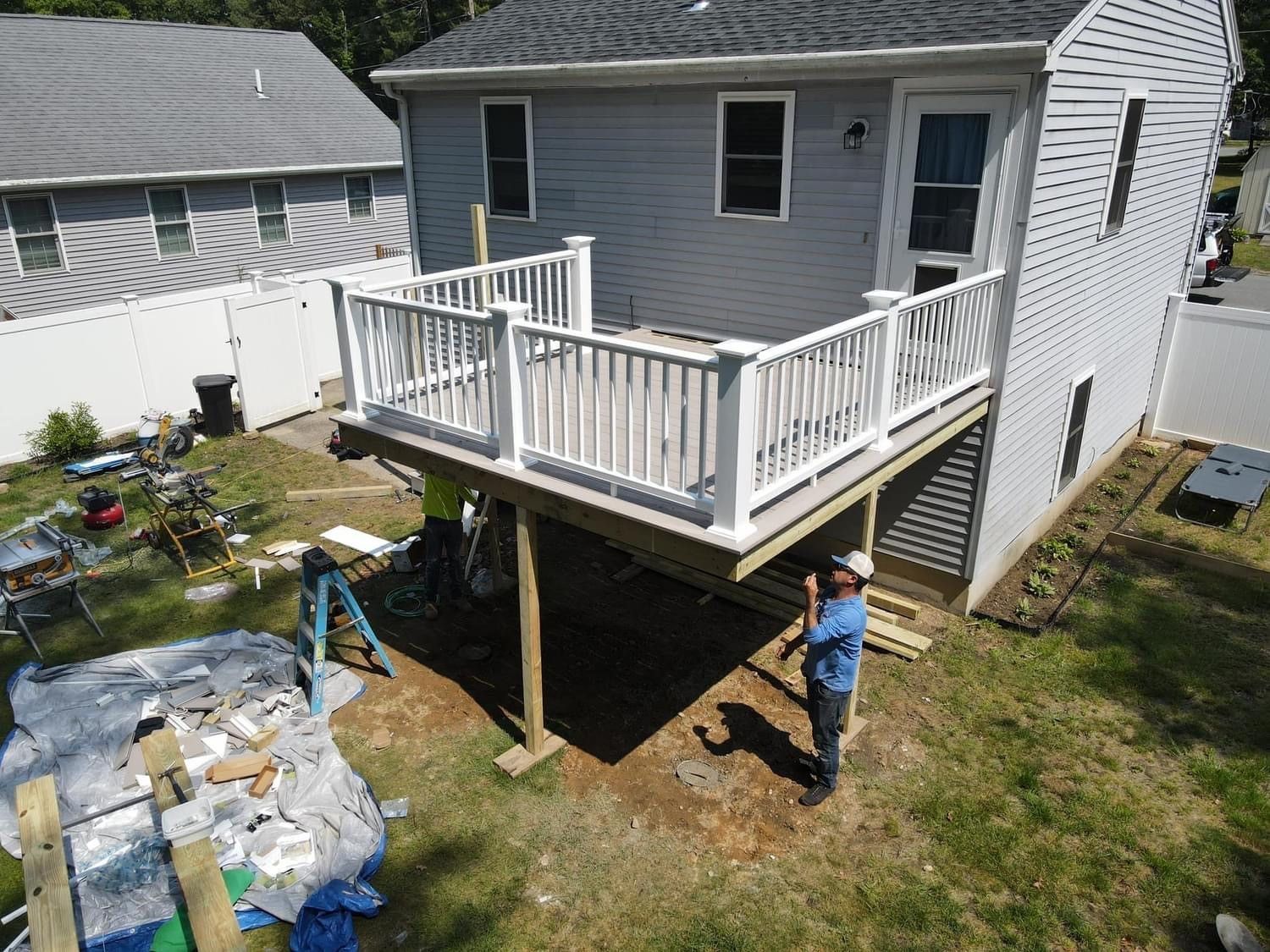 Backyard deck with white railing beside a house; a worker stands below near construction materials.
