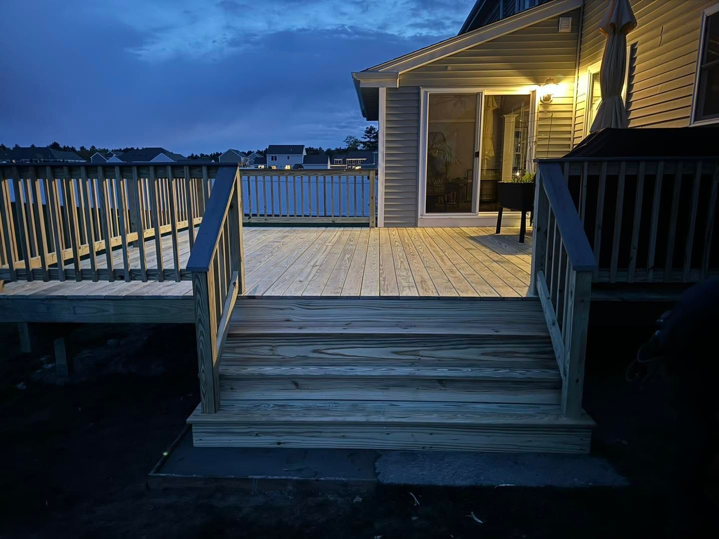 Wooden deck steps leading to a lit patio at dusk with a waterfront view beyond the railing