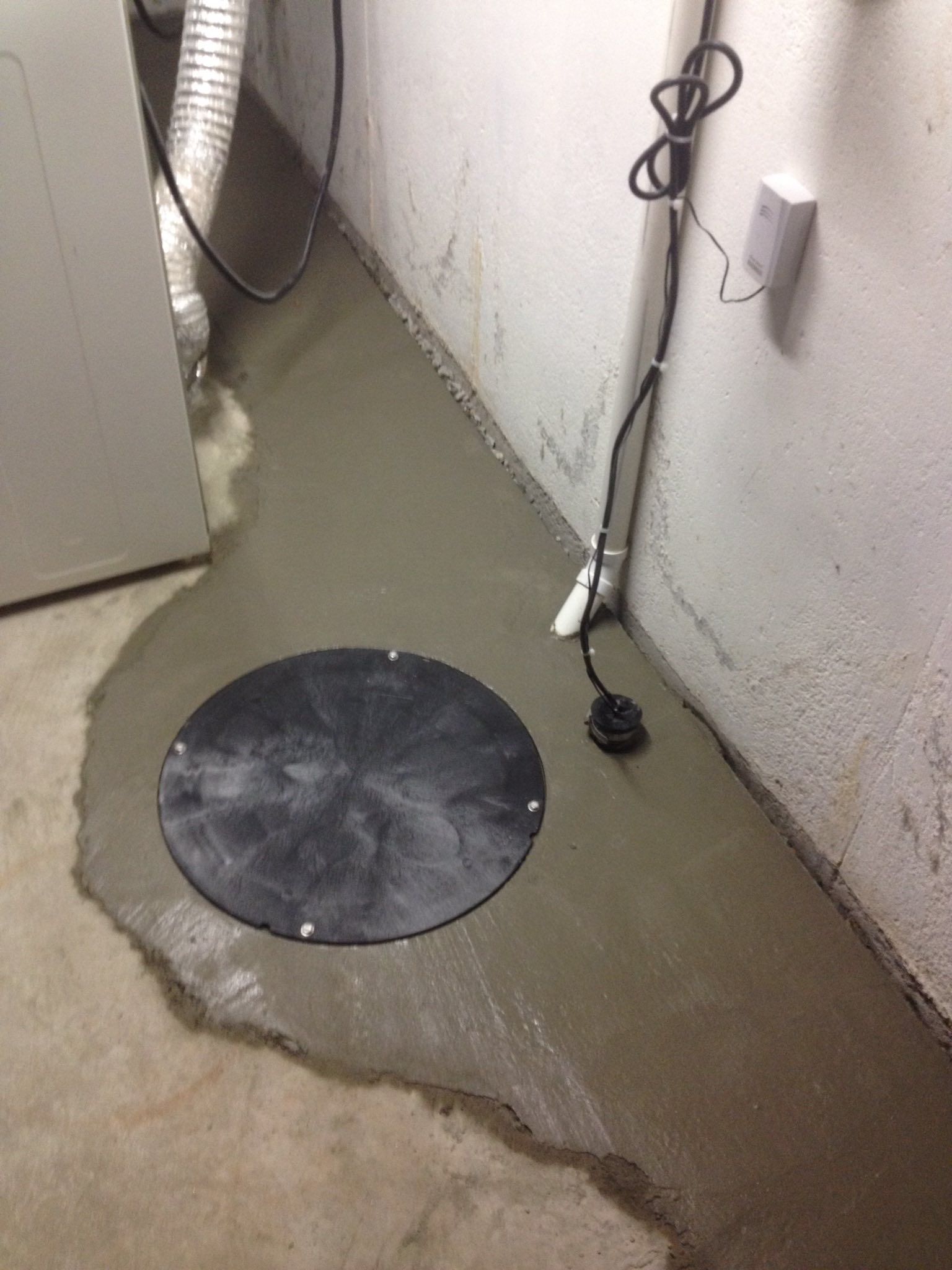Basement utility area with standing water, a round drain cover, and exposed pipes along the wall
