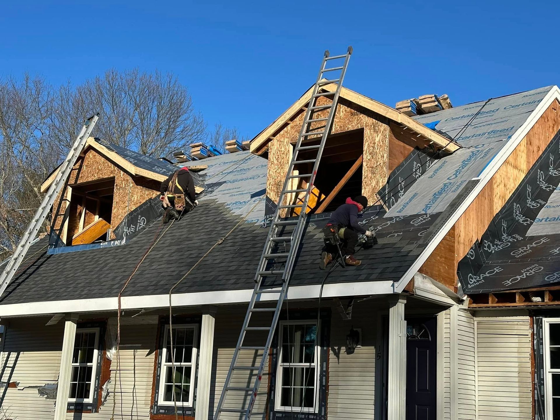 Workers repairing a house roof with ladders, exposed plywood, and dark shingles under a blue sky