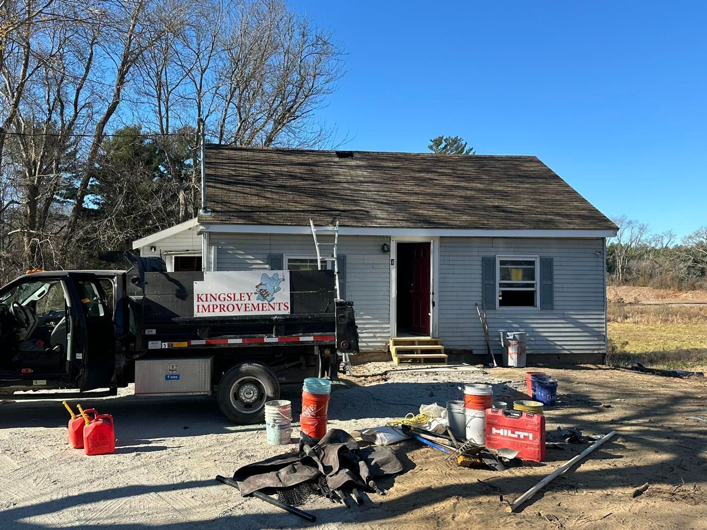 Work truck, tools, and red fuel cans outside a small gray house under repair