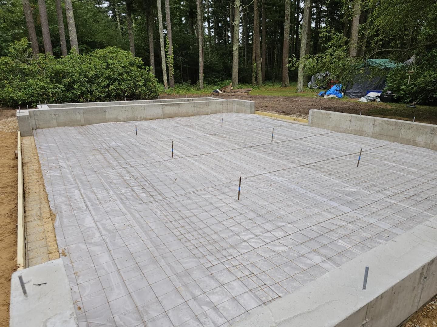Concrete slab foundation with rebar grid, surrounded by trees and dirt at a wooded construction site