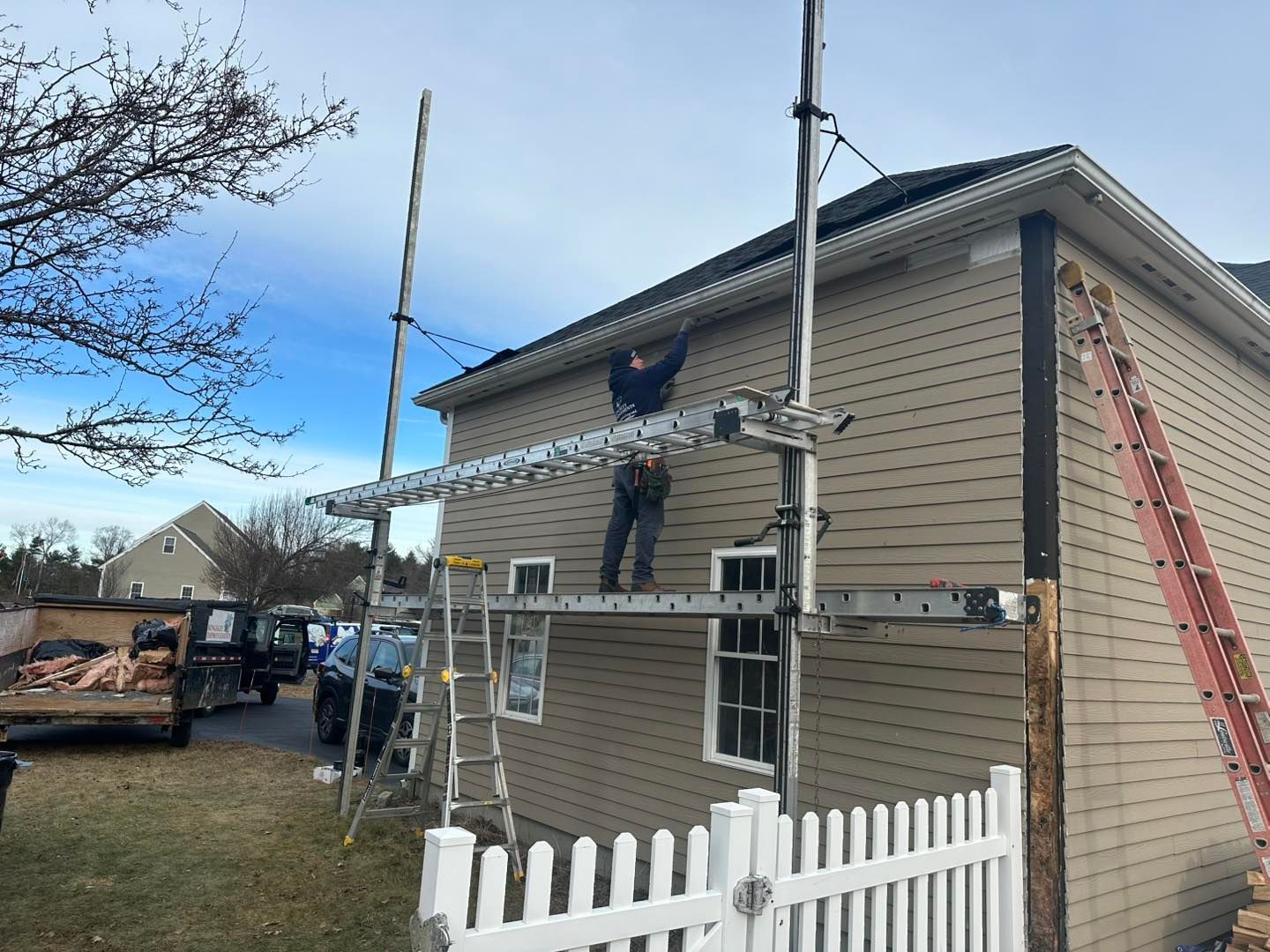 Workers on ladders repairing the siding and roof of a beige house with white fence