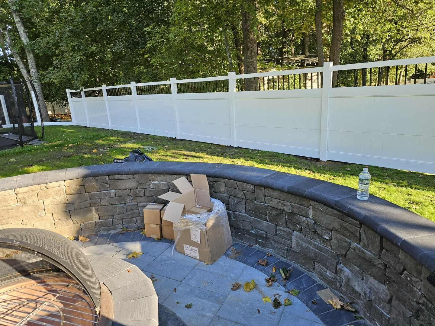 Stone fire pit with cardboard boxes on the floor and a white fence in a backyard.