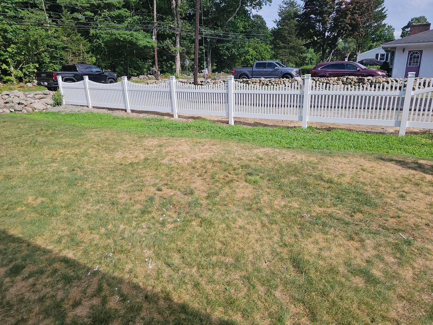 White picket fence along a grassy yard with parked cars and trees in the background.