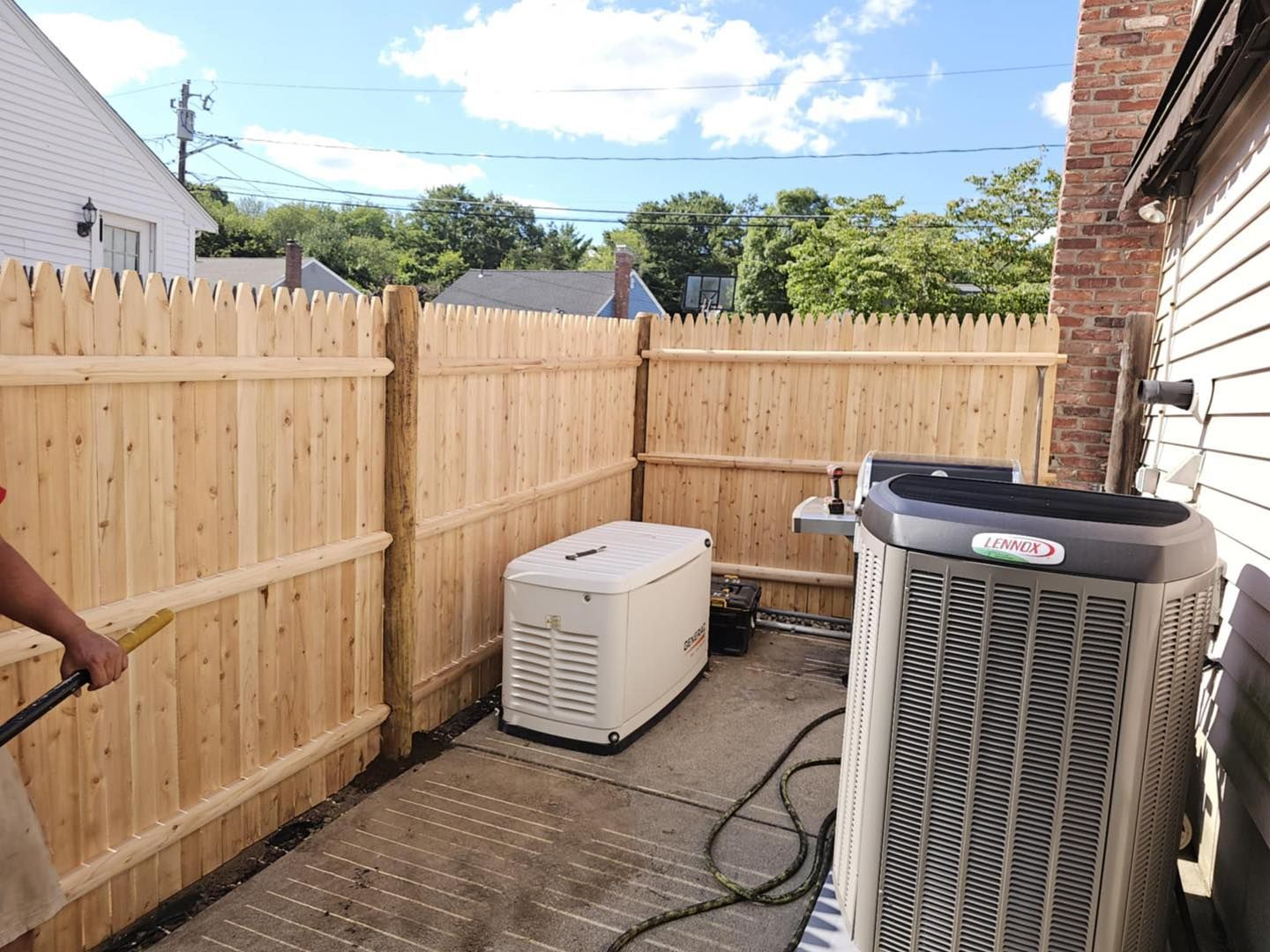 Backyard patio with two outdoor HVAC units beside a wooden privacy fence under a blue sky.
