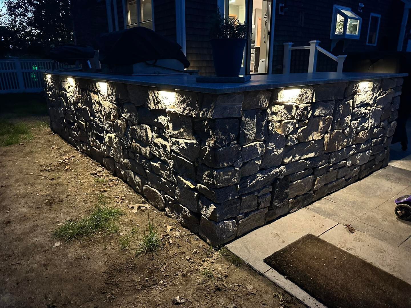 Stone retaining wall lit at night beside a dirt yard and concrete walkway with a doormat