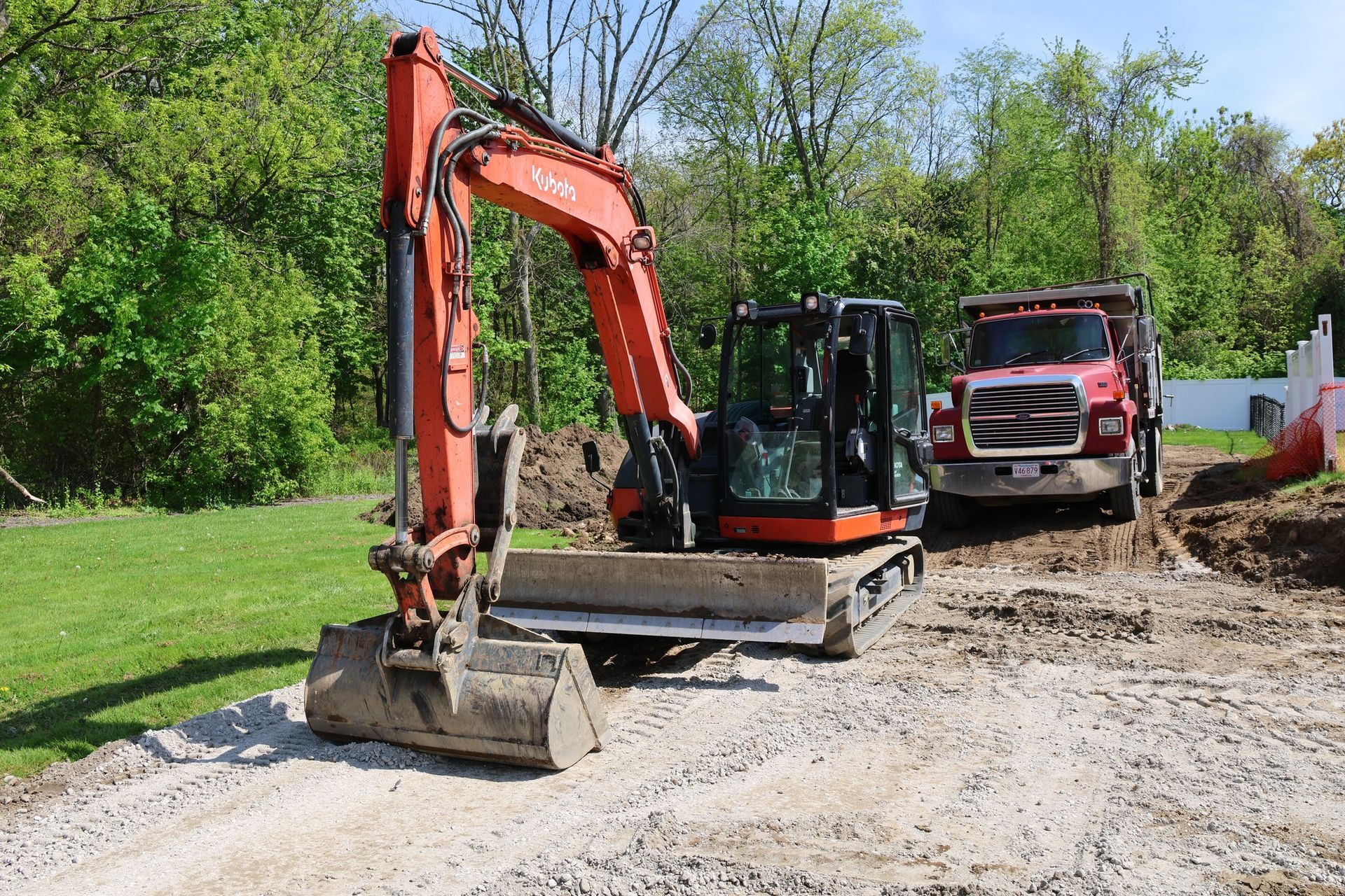 Orange excavator and red dump truck on a gravel path beside a grassy, tree-lined worksite