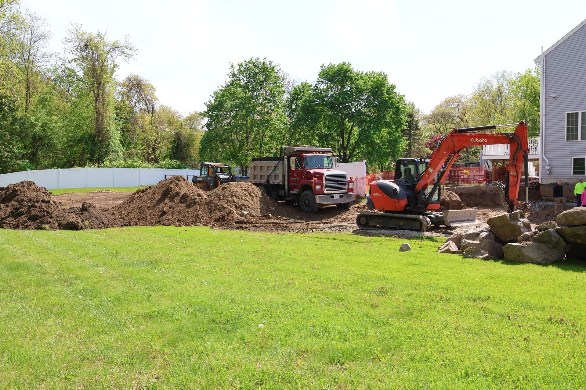Construction equipment working beside a house, with dirt piles and an excavator in a grassy yard.