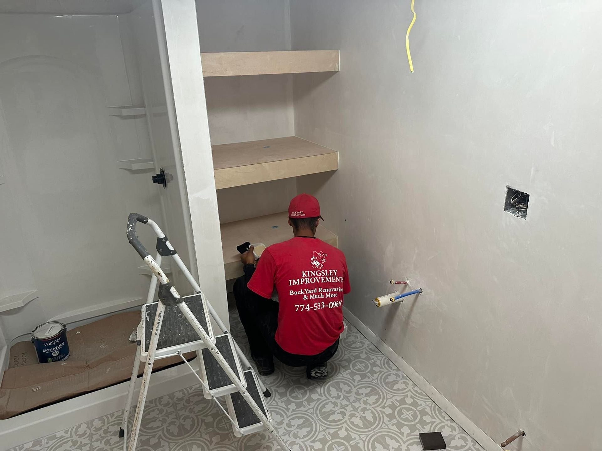 Worker in a red shirt installing shelving in a small room with a step ladder and tools.