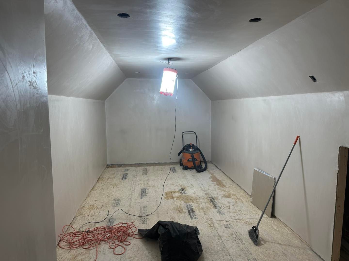 Empty unfinished room with sloped white walls, tools, extension cord, and shop vacuum on the floor.