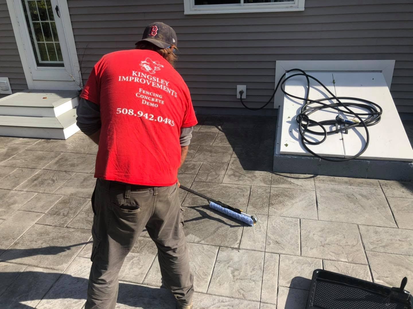 Worker in red shirt pressure washing a patio beside a house, with hose coiled on a white unit.