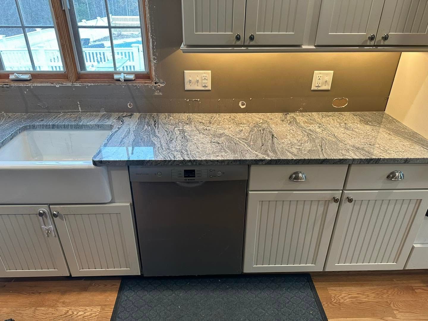 Kitchen countertop with white sink, dark dishwasher, and white cabinets under a window.