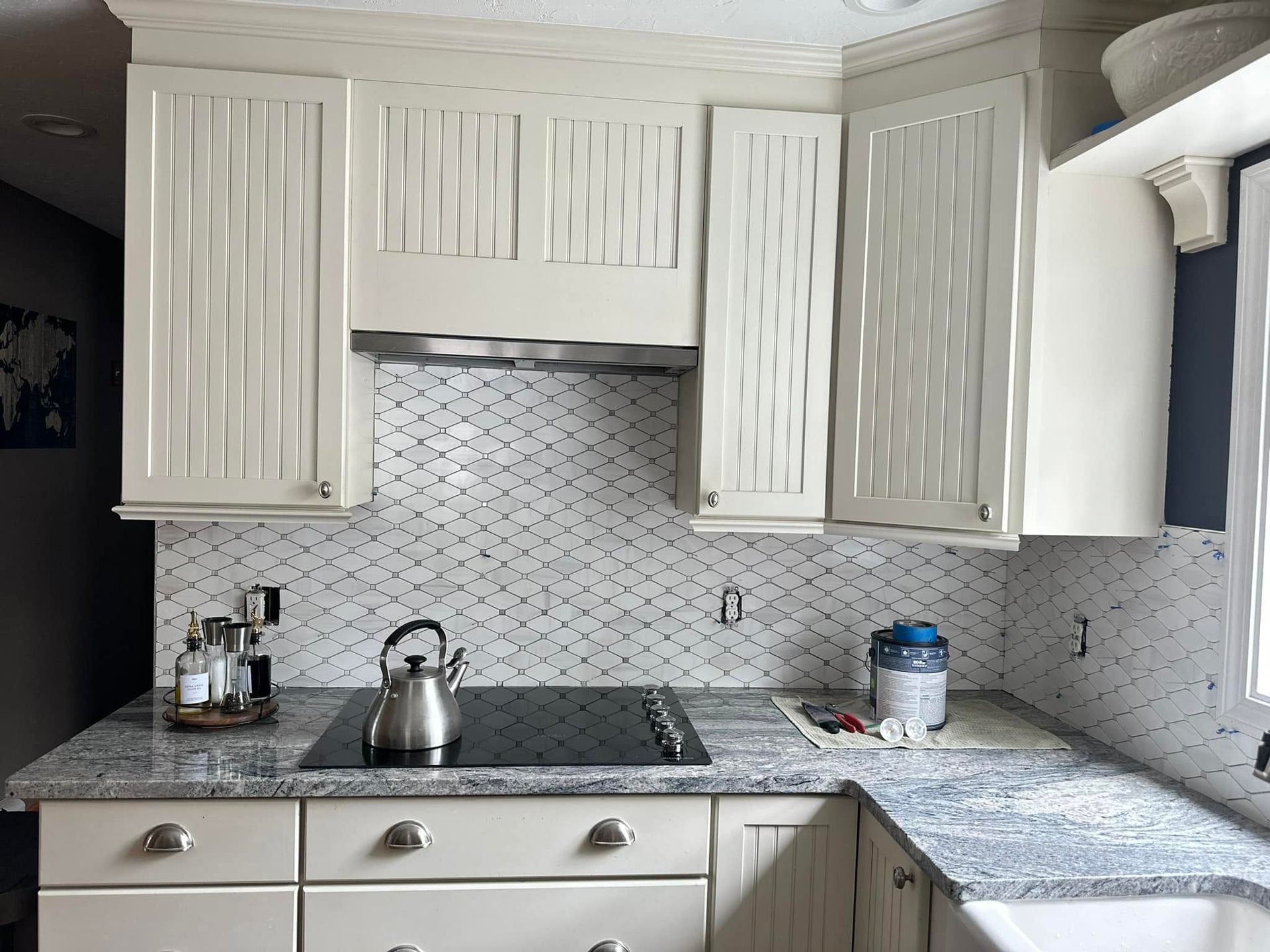 White kitchen with cabinets, black countertops, patterned tile backsplash, and a kettle on the stove.