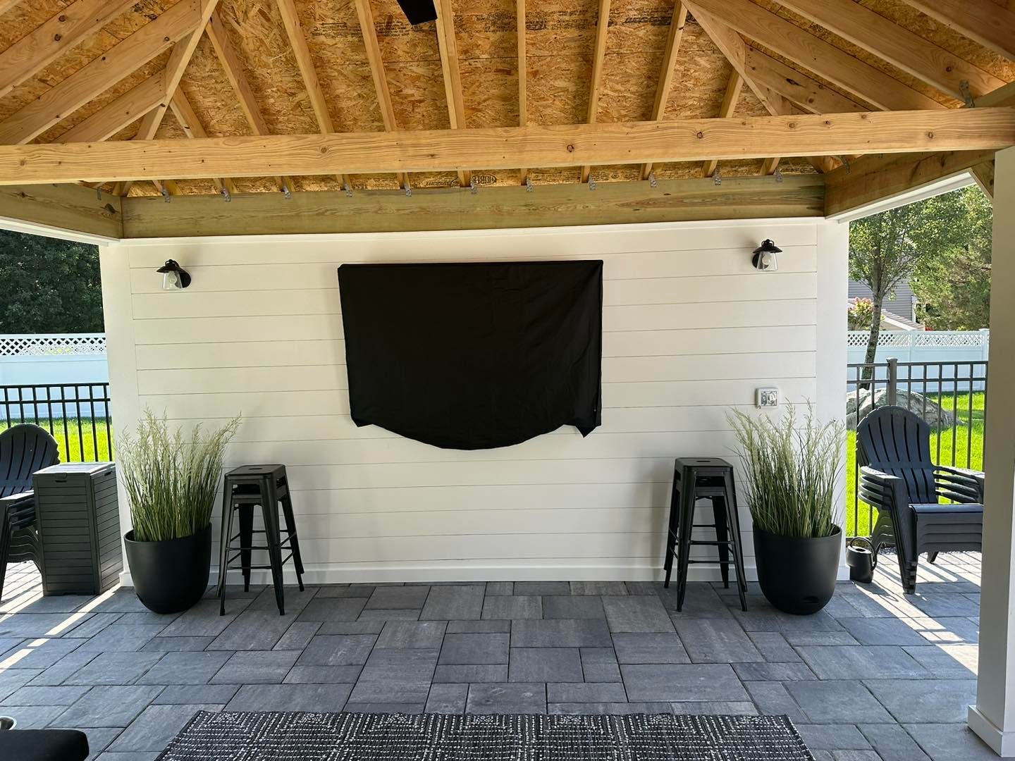 Outdoor bar pavilion with white wall, black drape, two stools, potted plants, and stone patio under a wooden roof