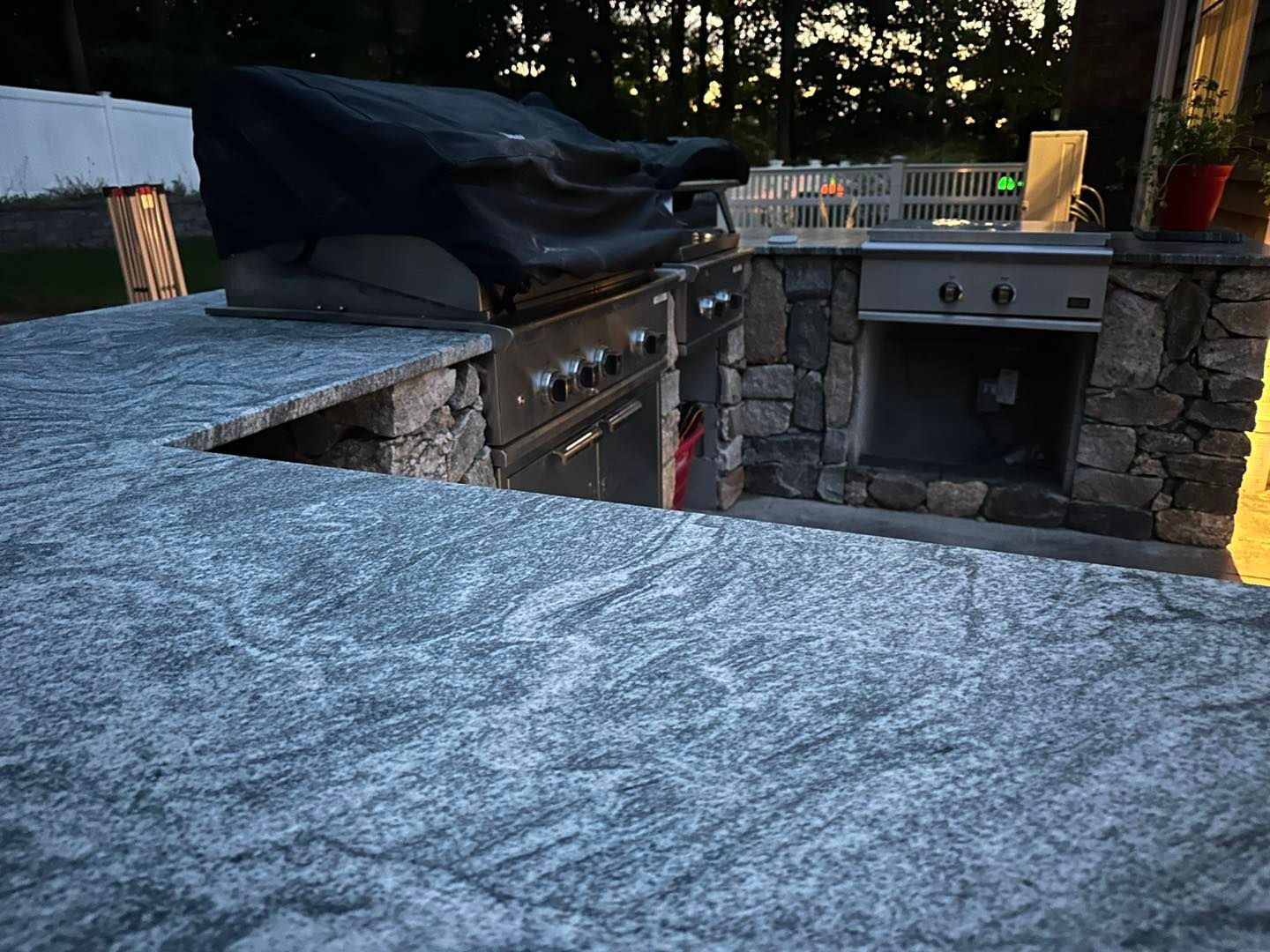 Outdoor stone patio kitchen with built-in grill, sink, and covered counter at dusk