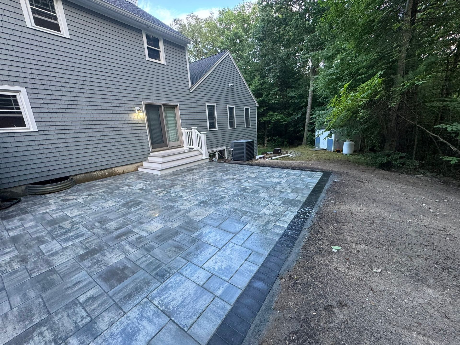 Backyard patio with gray pavers beside a gray house, bordered by gravel and trees