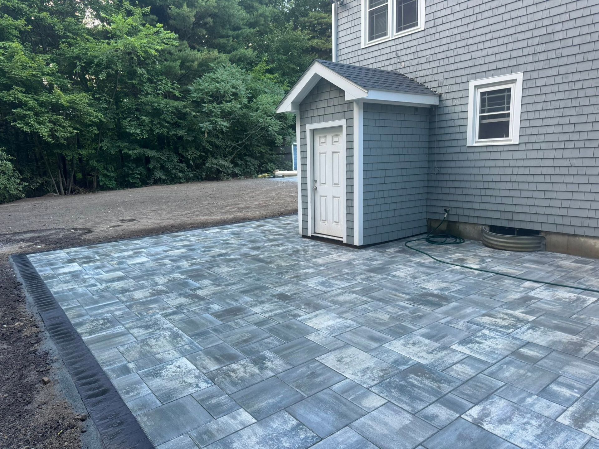 Paver patio beside a gray house with a small white shed and wooded background