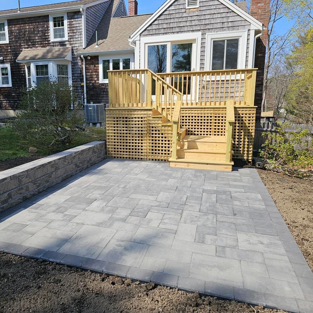Backyard patio with gray pavers leading to a wooden deck and steps beside a house