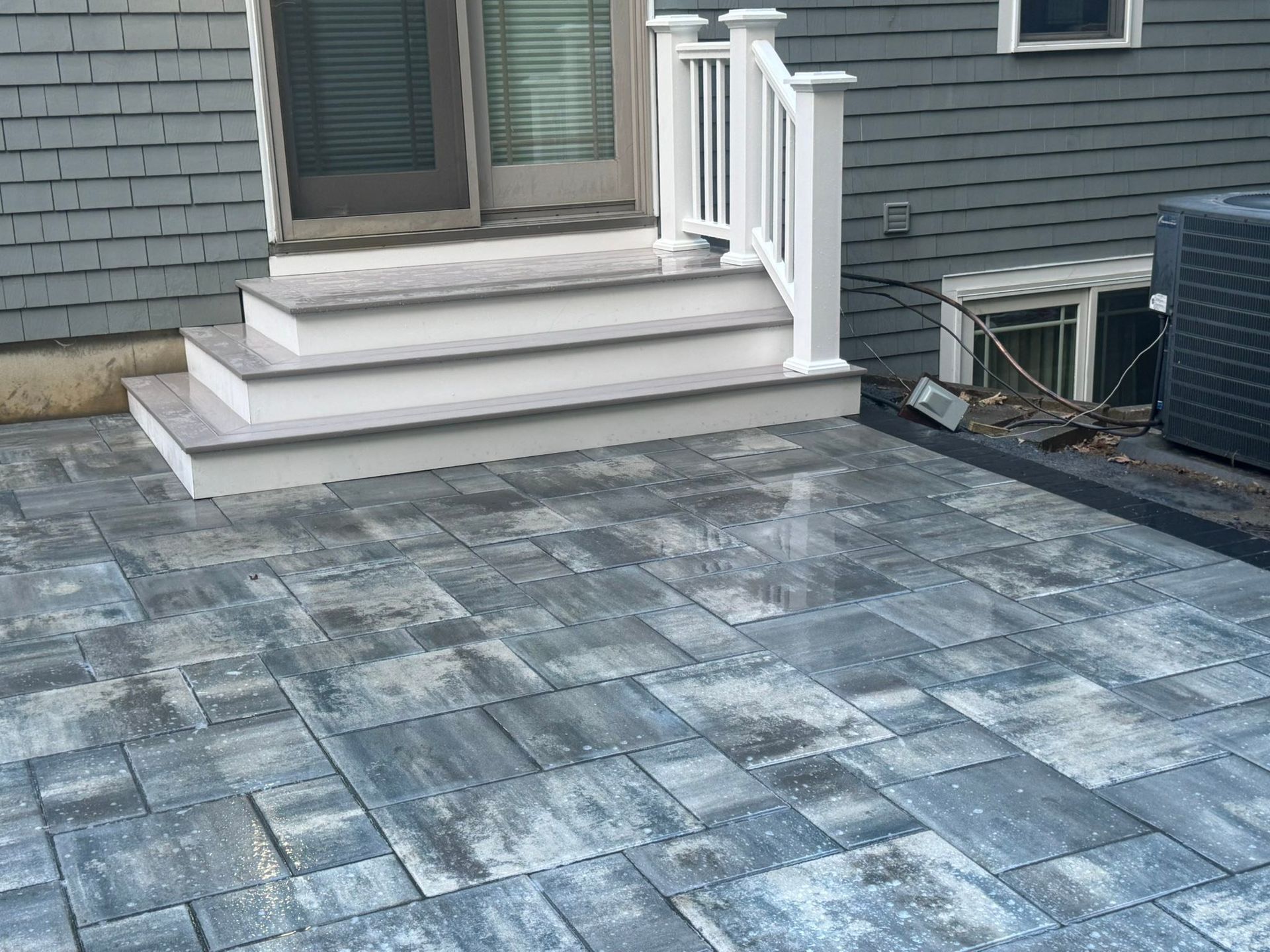 Paver patio beside gray house steps with white railing and a basement window well