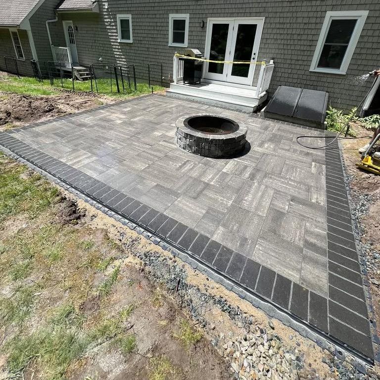 Backyard patio with gray pavers, circular fire pit, and black border beside a house and deck.