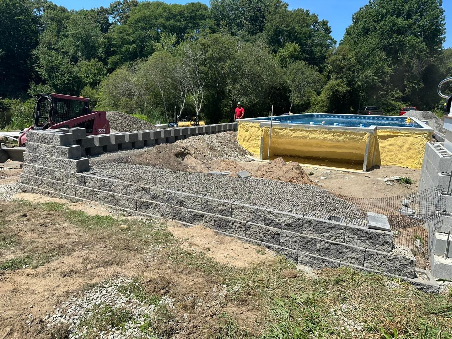 Construction site with a stone retaining wall, yellow-lined pool or pond, and workers in the background