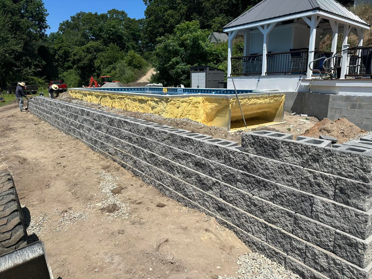 Stone retaining wall under construction beside a house, with dirt, tarp, and a porch in the background