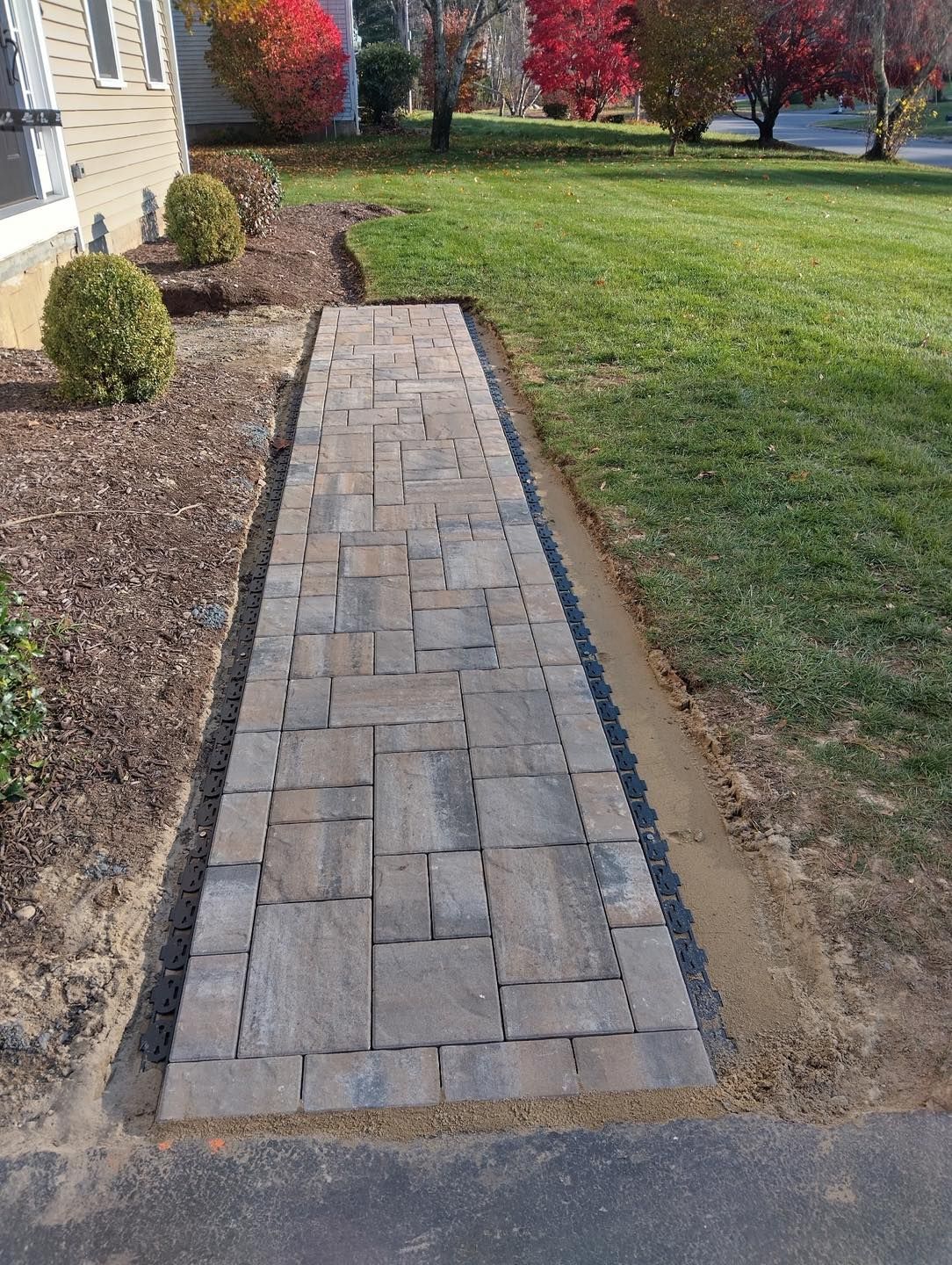 Narrow stone paver walkway beside a house, bordered by mulch and grass with autumn trees in the background