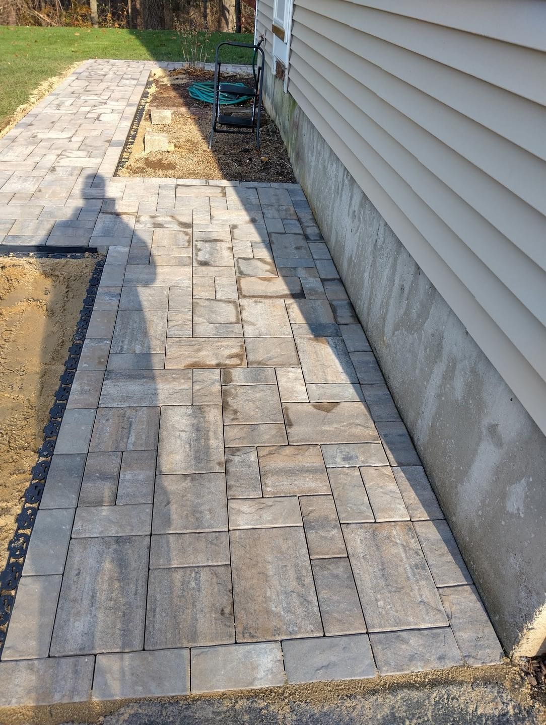 Stone walkway beside a house, with tan pavers and a small landscaped edge.