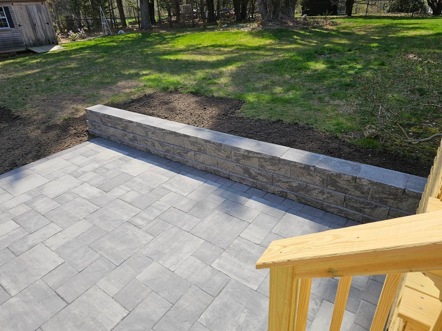 Paver patio beside a low stone retaining wall and grassy yard, with wooden steps in the foreground.